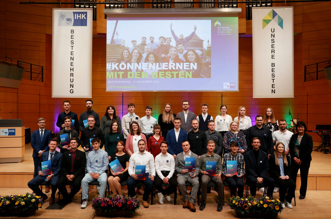 Gruppenfoto auf einer Bühne: junge Erwachsene mit Urkunden in der Hand; im Hintergrund Banner der IHK-Bestenehrung und eine Leinwand mit dem Motto „#KÖNNENLERNEN MIT DEN BESTEN“.