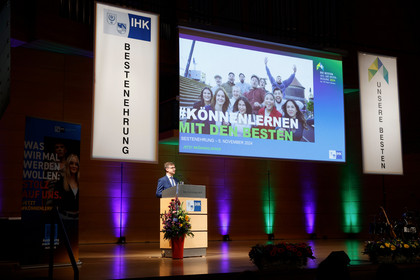 in Redner steht am Pult auf einer Bühne bei einer IHK-Veranstaltung zur Bestenehrung; hinter ihm eine große Leinwand mit dem Motto „#KÖNNENLERNEN MIT DEN BESTEN“ und bunte Beleuchtung an der Rückwand.