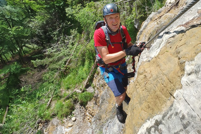 Eine Person in rotem T-Shirt und Helm klettert an einem gesicherten Felsabschnitt entlang, umgeben von dichtem Grün.