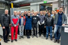 Eine Gruppe von Teilnehmenden steht in einer Werkhalle der SLV Halle für ein Gruppenfoto zusammen. Die Personen tragen Arbeitskleidung, Freizeitkleidung oder Schutzkleidung. Im Hintergrund sind industrielle Maschinen und Lüftungsanlagen sichtbar.