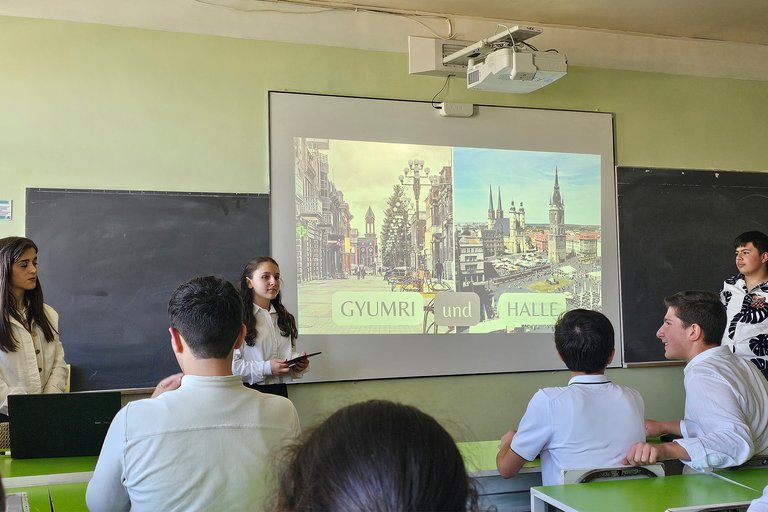 Eine Schülergruppe präsentiert eine PowerPoint-Folie mit dem Titel „GYUMRI und HALLE“ in einem Klassenzimmer.