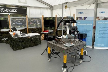 Messezelt mit mobiler Roboterzelle auf Schweißarbeitsplatte, im Hintergrund zwei SLV-Halle-Rollups und ein Stand mit Tarnnetz, Monitor und 3D-Druck-Beschilderung