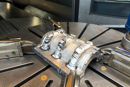 Metallisches Bauteil mit präzise aufgetragenen Schweißraupen ist auf einem runden Maschinentisch fixiert, Spannvorrichtungen halten das Werkstück in Position