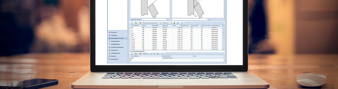 Ein geöffneter Laptop steht auf einem Holztisch, auf dem Bildschirm ist eine technische Software mit Schweißnahtdarstellungen und tabellarischen Daten zu sehen. Daneben liegen eine Computermaus und ein Smartphone, im Hintergrund ist eine Werkhalle unscharf erkennbar.