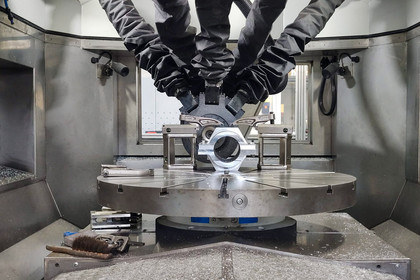 Innenansicht einer Fertigungsanlage mit mehreren flexiblen Roboterarmen über einem runden Maschinentisch, ein metallisches Werkstück ist eingespannt, Späne und Werkzeuge im Vordergrund