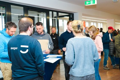 Personengruppen stehen in einem Veranstaltungsbereich und unterhalten sich. Informationsmaterial liegt auf Stehtischen.