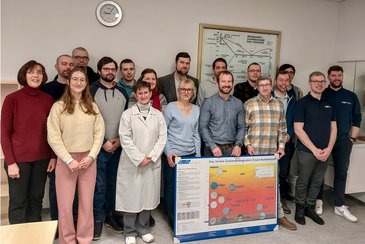 Die Teilnehmenden aus der posieren für ein Gruppenbild. Im Vordergrund ein als Gastgebergeschenk gestaltetes Eisen-Kohlenstoffdiagramm, im Hintergrund ein ehrwürdiges Exemplar aus den 1960er Jahren.