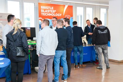 Mehrere Personen stehen an einem Ausstellerstand mit der Aufschrift „Bristle Blaster© Technology“ und führen Fachgespräche. Im Hintergrund weitere Informationsstände.