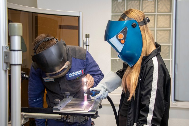 Mädchen mit Schutzhelm hält eine Schweißelektrode, die von einem Mitarbeiter in Schutzkleidung geführt wird.