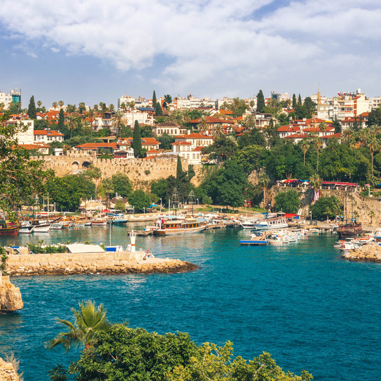 Blick auf den Hafen und die Altstadt von Antalya mit Booten im türkisblauen Wasser, umgeben von grünen Bäumen und hellen Häusern.