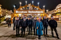 Eine Gruppe von Mitarbeitenden der SLV Halle GmbH und der SLV Hannover steht vor dem beleuchteten Eingang des Dresdner Striezelmarkts. Die Teilnehmenden tragen festliche Mützen und genießen die weihnachtliche Atmosphäre.