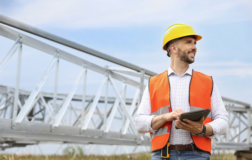 Mann mit Helm und Warnweste hält ein Tablet und steht vor einer Stahlkonstruktion im Freien, blauer Himmel im Hintergrund.