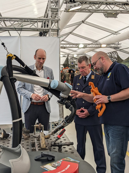 Drei Männer stehen an einer Schweißarbeitsplatte mit Roboterarm, einer im Bundeswehranzug, einer im Anzug und einer mit SLV-Halle-Poloshirt hält ein orangefarbenes Werkstück