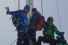 Drei Bergsteiger stehen auf einem Gipfel vor einem Gipfelkreuz. Sie tragen warme Outdoor-Kleidung mit Kapuzen und Rucksäcken. Das Wetter ist neblig, und sie lächeln in die Kamera.