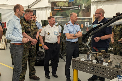 Mehrere Männer, darunter uniformierte Bundeswehrangehörige, stehen in einem Ausstellungszelt vor einem Roboterarm an einem Schweißtisch.